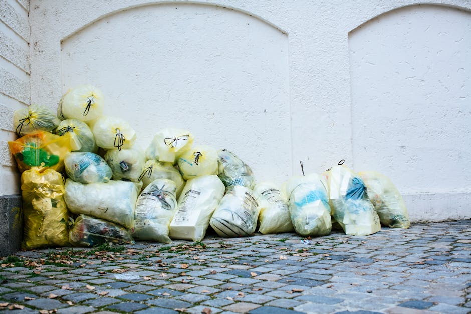 A large pile of black rubbish bags stacked against a modern tiled wall, with some bags torn open revealing plastic bottles and paper waste inside. The bags are situated outdoors on a paved surface, an