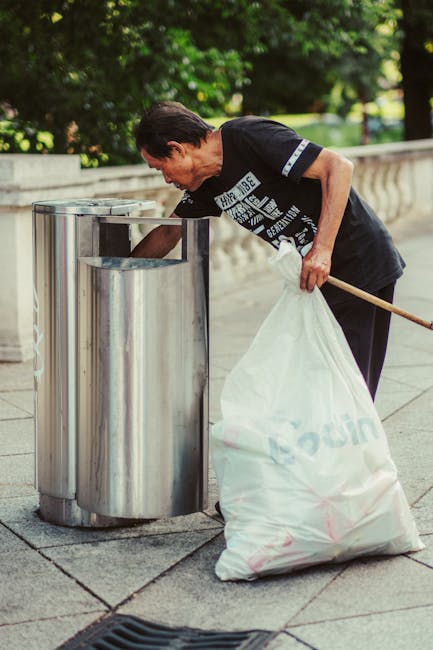 A middle-aged man with short dark hair, dressed in a black t-shirt with white graphic text on the front, is seen disposing of waste into a stainless steel litter bin situated on a paved outdoor area.
